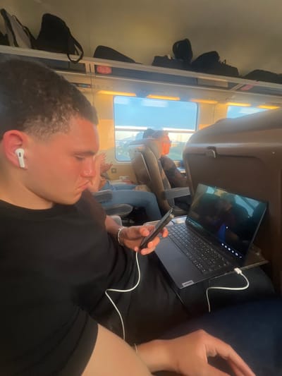 The image shows a young man sitting on a train in Blois, France (latitude: 47.594, longitude: 1.324). He is wearing a black T-shirt and blue jeans. He is holding a smartphone in his right hand and is looking at it. A white wire from a white earbud in his left ear leads to the smartphone. A black laptop computer with a bright screen is on the seat beside him. To the right of the man, an older man is sitting in a seat facing a large window. The train is a modern, clean vehicle. There are overhead luggage racks with luggage.  The image was taken from the perspective of someone sitting across from the young man.  The sun is shining brightly outside. The man sitting by the window is looking out the window.  The scene is inside a train, and the environment is a modern train car.