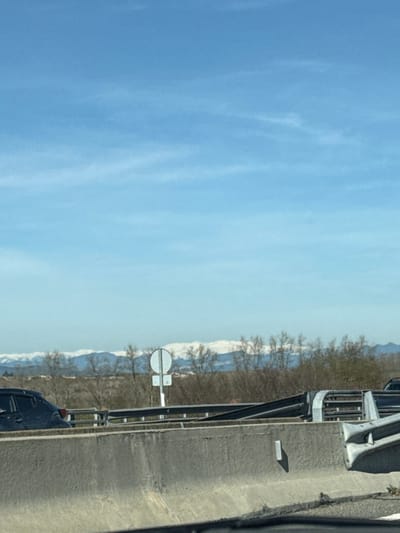 The image is a landscape photograph, appearing to be taken from a vehicle, with a clear sky, distant snow-capped mountains, and a roadside scene. The image is originally oriented vertically but depicts a horizontal vista.

The upper portion of the frame is dominated by a bright, clear blue sky with subtle, wispy white clouds, suggesting a sunny day. Below the sky, a distant horizon reveals a line of mountains, their peaks and upper slopes covered in white snow, indicating a cold climate or high altitude. In front of the mountains, a band of bare, deciduous trees or shrubs with dark branches stretches across the midground, suggesting late winter or early spring.

The lower part of the image depicts a road or highway environment. A grey, possibly concrete or asphalt, road surface is visible, along with a dark, metal barrier or guardrail running parallel to the road. Near the barrier, approximately in the lower-middle portion of the scene (when correctly oriented), there is a white circular road sign with a thick horizontal black bar in its center, typically indicating "No Entry" or "Do Not Enter" for vehicles. A smaller, rectangular sign is attached directly below it, but its details are indistinguishable. Further to the right, parts of vehicles are visible, including the front quarter of a light-colored (possibly white or silver) truck or van and a darker vehicle further back, indicating the perspective is from within or alongside moving traffic. A distinct, rectangular shadow is cast on the road surface, suggesting sunlight from the side.

The overall scene conveys a sense of travel through a scenic, mountainous region. The time of day appears to be bright daylight, and the presence of snow on the mountains coupled with bare trees suggests the season is late winter or early spring. No discernible text is visible on any signs or objects.