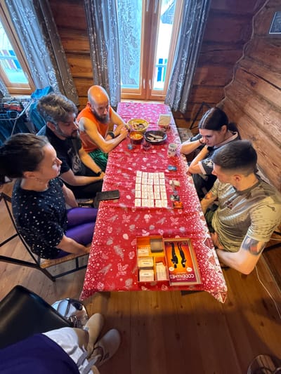 Six individuals are present indoors, seated around a wooden table covered with a red tablecloth featuring white leaf patterns, in what appears to be a cabin or wooden dwelling located in the city of Oshutyaly, Russia. The room features exposed log walls and a wooden floor. Two large windows with patterned curtains are visible at the far end of the table, showing an exterior view.
From left to right: A person is partially visible, only their legs and feet in light grey pants and tan slippers are seen. Next, a woman with dark hair in a black polka-dotted top and bright purple leggings sits on a wicker chair, observing the table. Adjacent to her is a man with grey hair and a beard, wearing a black t-shirt, also looking towards the table. Directly across from the first woman, a bald man with a beard, wearing an orange tank top and patterned shorts, leans forward intently. Next to him, a woman with dark hair and a dark top with white detailing rests her arm on the table, gazing downwards. Lastly, a man with short dark hair and a patterned short-sleeved t-shirt with a tattoo on his left forearm is seated, directing his attention to the table's contents.
The central activity involves a game of "Codenames." The game box is open on the lower right of the table, displaying components. A grid of approximately 25 white word cards is laid out in the center of the table, surrounded by smaller game key cards and standees. Various snacks are arranged in bowls at the upper end of the table, including what appears to be chips, candy, and nuts. A white mug with "ARAN" visible and a clear glass with blue liquid are also on the table. A black smartphone is placed on the tablecloth near the word cards. The individuals appear engaged in playing the board game in a casual setting.