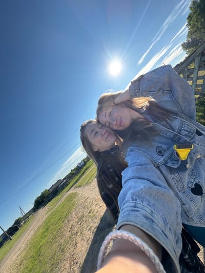 The image depicts an outdoor selfie of two young women in Ust'-Silayka, Russia, taken on a bright, sunny day. The sky is a clear blue with sparse, wispy white clouds and a prominent sun radiating lens flare from the upper right quadrant.

The woman closer to the camera, positioned on the right, has light brown hair and a smiling expression with her head slightly tilted. Her right arm is raised, with her hand near her hair. She is wearing a light-wash denim jacket featuring a black heart patch and embroidered text on the left chest area, and a yellow, multi-pronged object, possibly a pet grooming tool, is clipped to her right jacket pocket. A white braided bracelet is visible on her left wrist. The second woman, positioned slightly behind and to the left, also has light brown hair, closed eyes, and a smiling expression, with her head leaning against the first woman's. She exhibits visible freckles on her nose and cheeks.

The background reveals a rural or semi-rural environment. A dirt path or road with interspersed patches of green grass extends into the distance on the left side. Several wooden utility poles with overhead power lines are visible along this path. In the middle ground on the left, multiple simple, wooden structures or houses are present. Trees are scattered throughout the background, and a wooden lattice fence is partially visible on the far right. The photograph has a tilted perspective.