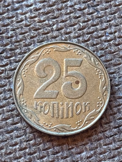 This is a close-up photograph of a 25 Kopek coin from Ukraine, resting on a textured, woven surface. The coin is the central subject, with its metallic sheen and embossed details clearly visible. The coin features a large "25" in the center, with the numeral "2" prominently displayed below a smaller "5". To the right of the numerals, the word "КОПІЙОК" (Kopek) is inscribed in Cyrillic. Decorative elements, including small circles resembling berries and stylized leaves, form a border around the coin's edge. A small dark spot, possibly a blemish or part of the design, is visible within the upper loop of the "5". The coin is placed on a dark, rough fabric that appears to be a knitted or woven material, possibly a blanket or upholstery. The lighting suggests an indoor setting, with shadows cast by the coin onto the fabric, indicating the light source is from the side and slightly above. There are no people or explicit events depicted; the image focuses solely on the coin and its immediate surroundings. The overall impression is one of still life photography, highlighting the details of the currency. The context provided indicates the coin is from Pereiaslavske, Ukraine, but this specific location is not visually discernible from the image itself.