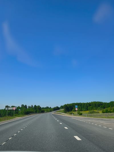 The image shows an asphalt road curving to the right, bordered by a metal guardrail on the left and a grassy shoulder with trees in the background. The sky is clear blue with faint, wispy clouds. There are two traffic signs visible on the left side of the road. The first sign, a circular red and white sign, depicts two cars side-by-side with a red bar through them, indicating no overtaking. The second sign, a blue square sign, shows a left turn arrow with a straight arrow to its right. The scene appears to be in a rural or suburban area, possibly on the outskirts of Makaryev, Russia, given the presence of trees and the general landscape. There are no people or vehicles visible in the immediate vicinity of the road.