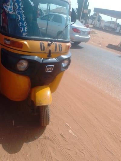 An image captured in Lapai, Nigeria, depicts the front left quarter of a yellow and black Bajaj RE auto-rickshaw, parked on a reddish-brown dirt surface adjacent to a paved road. The auto-rickshaw's front features circular headlights, a black bumper and cowl, and a "BAJAJ RE" emblem. The number "676" is painted in black on the yellow cowl above the windshield. A blue and white patterned fabric is visible through the top left of the windshield. A silver sedan (likely a Toyota Corolla) is partially visible in the reflection of the auto-rickshaw's windshield and positioned directly behind it on the paved road, facing away. In the distant background on the right, a fuel station with visible pumps and signage is present. Trees line the horizon under a bright, clear sky. The ground in the immediate foreground shows multiple tire tracks in the reddish-brown earth. No individuals are clearly discernible.