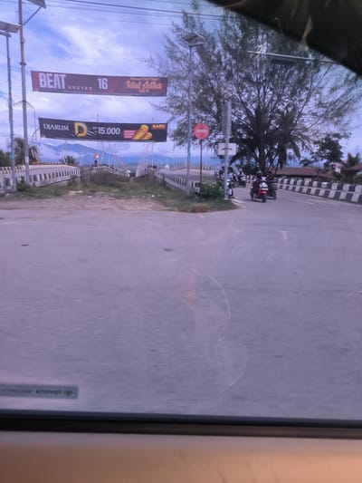 Captured from inside a vehicle, this image presents a view of a road junction or intersection in Paya Tringgadeng, Indonesia, with prominent banners overhead. The sky is overcast, suggesting either an approaching evening or a cloudy day.

In the upper part of the frame, two large banners are suspended across the road. The top banner advertises "BEAT KRETEK 16" and extends "Selamat Hari Raya Idul Adha 1447 H," indicating a celebratory period. Below it, a black banner for "DJARUM D" cigarette brand displays a price of "Rp 15.000" and the word "BARU" (New), along with an illustration of cigarette sticks. Utility poles with solar panels are visible, supporting these banners.

The road itself is concrete or asphalt, with multiple lanes. To the left and right, parts of bridges or elevated roadways with concrete barriers featuring white and black stripes are visible. An Indonesian flag stands on one of the bridges in the distance, and mountains frame the background. A large, dense-foliage tree stands prominently on the right side of the road, partially obscuring the view beyond it. Beneath the tree, a red circular "no entry" sign is visible.

Several individuals are seen riding motorcycles on the right side of the road, heading away from the camera. The foreground of the image shows reflections on a car's windshield or dashboard, including partially visible text like "ATOYOT" (likely a reversed "TOYOTA" logo). The scene depicts a busy, yet somewhat rural, setting during a festive season.