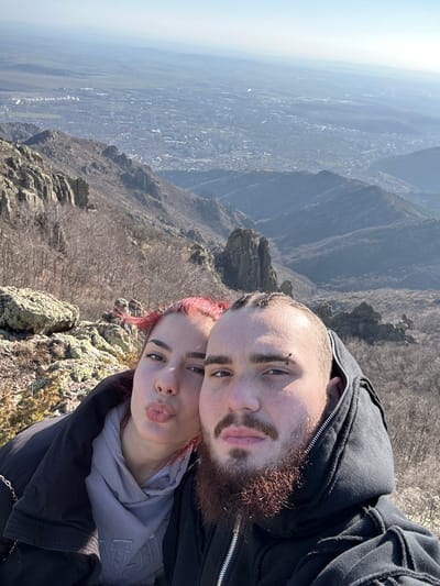 The image depicts two individuals, a male and a female, in a close-up, selfie-style photograph taken on a mountainous elevation overlooking the city of Ichera, Bulgaria. The background reveals a wide urban expanse under a clear, bright sky.
The female, positioned to the left, has red hair and is dressed in a dark jacket over a light grey hoodie. Her lips are pursed. The male, positioned to the right, displays braided hair on the frontal scalp, a full brown beard, and a piercing above his left eyebrow. He wears a dark, zippered hooded sweatshirt. His expression is neutral.
The immediate environment consists of rugged, rocky terrain with sparse, bare deciduous trees, indicative of late autumn, winter, or early spring conditions. Far below, the city extends into the valley, bordered by further distant mountain ranges under a slightly hazy horizon.