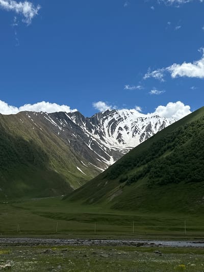 A wide-angle, full-shot photograph captures a majestic mountain landscape under a clear blue sky with scattered white clouds. The image is dominated by a mountain range, with the peaks covered in snow, contrasting with the lush green slopes. The foreground features a grassy field dotted with small white and yellow flowers, with a rocky stream flowing through it. In the midground, a vast green valley stretches out, with a few utility poles sparsely distributed across it. The scene conveys a sense of natural beauty and tranquility, likely depicting the scenery around Ketrisi, Georgia, a region known for its stunning mountain vistas. There are no people or animals visible in the frame.