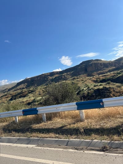 This media file depicts a roadside scene with a mountainous backdrop. The primary subjects include a metal guardrail with blue and white markings, a paved road, dry grass, and a steep, rocky mountainside. The scene is set on the side of a road, likely in a rural or mountainous area.

The image appears to have been taken during the daytime, under clear, blue skies with a few scattered white clouds. The lighting suggests it is a sunny day. The dry grass and the nature of the vegetation on the mountain suggest a warm or arid climate, possibly late summer or early autumn.

Notable details include the texture of the mountainside, which shows layers of rock and sparse vegetation, and the condition of the road, which appears to have some cracks and debris along the edge. The guardrail is a prominent object, acting as a visual separator between the road and the embankment.

There is no visible text in the image. The overall mood is neutral, focusing on the natural landscape and man-made infrastructure. Given the location context of Vayk, Armenia, it's reasonable to infer this is a scenic view along a road in that region, characterized by mountainous terrain. The image captures a slice of a journey, likely by vehicle, given the roadside perspective.