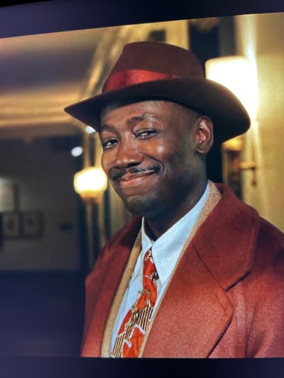 A close-up shot shows a Black man wearing a red fedora with a red band, a red coat, and a white shirt with a patterned tie. He has a mustache and is looking to his right with a slight smile. The background is blurred and appears to be an indoor setting with warm lighting and two framed pictures on the wall. The scene is a studio portrait.