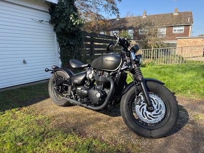 A matte black Triumph Bobber motorcycle is parked on a light brown gravel surface, adjacent to a green grass lawn, under direct sunlight in Babergh, United Kingdom. The motorcycle is oriented with its front wheel angled slightly to the right, presenting its left side. Key features include a black engine, black dual exhaust pipes, a single ribbed black seat, and black spoked wheels. The front wheel is equipped with a silver disc brake rotor displaying a spiral cut pattern. In the immediate background to the left, a white horizontally paneled garage door with a black handle is partially visible, with dense green ivy growing vertically on the wall beside it. To the right of the motorcycle, a wooden slat fence runs horizontally, separating the foreground from a more distant grassy garden area. Beyond the fence, two two-story residential buildings with brown brick facades, white-framed windows, and brown tiled roofs are discernible. Bare tree branches are visible above the fence line, and a low brick structure stands on the far right. No individuals or specific actions are observed in the scene.