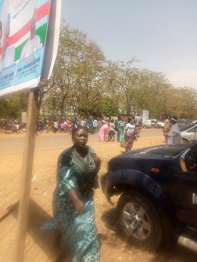 The image depicts an outdoor scene in Kagoma, Nigeria, under bright daylight conditions with a clear sky. The ground is dry and dusty.
In the immediate foreground on the left, a large political banner is partially visible, attached to a rough wooden pole. The banner features images of two men and text that includes "4+4=8," "Support Continued," "DR. SHEHU SARKI," and "HON. JEROME SILAS," set against a background of green, white, and red stripes.
A woman is prominently positioned slightly right of the frame's center. She wears a teal-green patterned traditional dress with long sleeves and a V-neckline. Her dark hair is pulled back, and she holds a blue object, consistent with a mobile phone, in her left hand. She is looking towards the camera. Immediately to her right, the front-right section of a dark blue vehicle, appearing to be an SUV or pickup truck, is visible, showing a silver-colored wheel rim and a portion of "IG" or "IGE" stenciled on the hood.
In the midground, a paved road extends horizontally across the frame. Beyond the road, a significant gathering of individuals is visible, appearing to be a crowd of people standing, walking, and interacting. Their clothing displays a variety of colors and patterns. Several motorcycles are present within this gathering and parked along the road. Further in the background, numerous additional vehicles, including cars and vans, are parked alongside the road. The background beyond the road and crowd consists of a dense line of trees, many with sparse foliage, extending across the horizon.