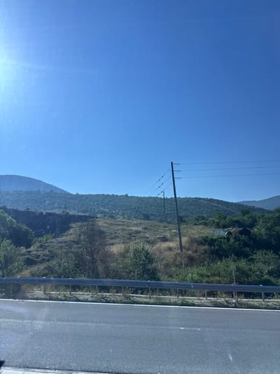 The image captures a rural landscape under a clear blue sky, suggesting an outdoor setting possibly in Kato Nevrokopi, Greece. The foreground features a paved road, likely a highway, with a metal guardrail separating it from the terrain beyond. The terrain itself is a mix of dry grasses, sparse trees, and rocky outcrops, with rolling hills covered in dense green foliage receding into the background. Several utility poles with power lines are visible, indicating the presence of infrastructure in the area. In the mid-ground, there are signs of human habitation or past structures, including what appear to be dilapidated sheds or outbuildings partially obscured by vegetation. The overall impression is one of a natural, somewhat wild environment with evidence of human presence.