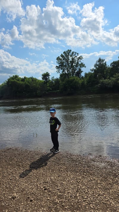 A young boy wearing a blue baseball cap, a black t-shirt, and black pants stands on a rocky shore beside a wide river. The boy is facing slightly away from the camera, with his right hand on his hip. The river's surface is rippled, and a line of green trees forms the opposite bank. The sky above is blue with scattered white clouds. The scene appears to be in an outdoor, natural setting, possibly near the village of Darino, Russia, given the overall landscape and vegetation.