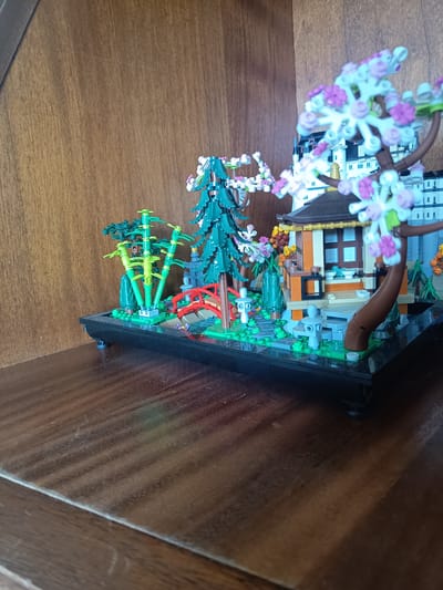 The image shows a detailed Lego model of a Japanese garden. The model features a small building with an orange facade and a dark roof, surrounded by greenery, including bamboo stalks and trees with pink and white blossoms. A red arched bridge crosses a small stream. The entire model is set on a black base with small feet. The background is a wooden surface, likely a shelf. There are no people or animals visible in the image. The overall scene is static and appears to be an indoor display. The city of Millstreet, Ireland is not depicted or referenced in this image.