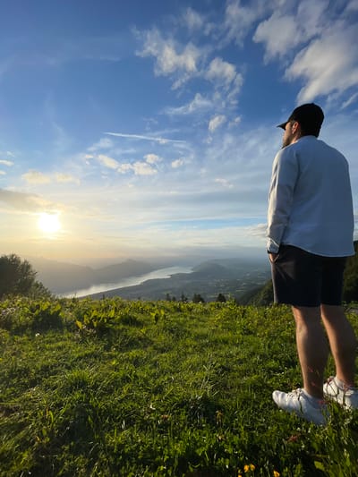 The image displays a male individual standing on a verdant hillside, gazing over a vast landscape in Les Déserts, France. The man, positioned with his back mostly towards the camera and slightly to the right, wears a black baseball cap, a light-colored long-sleeved shirt, dark shorts, and white sneakers. His hands are in his pockets, and his head is turned towards the horizon.
The foreground is comprised of dense green grass, dotted with broad-leafed plants and small yellow flowers, brightly illuminated by direct sunlight. Beyond the hillside, a wide body of water, resembling a lake or river, winds through a valley. This waterway is bordered by multiple layers of mountains and hills that recede into the distance, appearing hazy. A cluster of trees is visible on the left side of the frame, partially obstructing the view of the water.
The sun, a bright golden orb, is situated low on the horizon on the left side of the image, casting a warm, luminous glow across the entire scene and the lower sky. The upper sky is blue with scattered white cumulus clouds. A thin white contrail stretches horizontally across the mid-right portion of the sky. The lighting conditions are consistent with either sunrise or sunset.