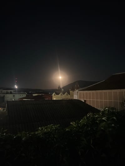 The image shows a night scene over a city, with a bright full moon in the sky. The moon is the dominant light source, creating a bright glow and lens flare. In the foreground, dark foliage is visible. Behind the foliage, a dark-tiled roof of a building is prominent. Beyond that, there are several other buildings with red and tiled roofs, as well as some modern structures with white and beige facades. In the background, hills or mountains are visible against the night sky. A tall radio tower with red lights can be seen on the left side of the image. A church spire is visible to the right of the moon. The overall impression is a quiet, illuminated urban landscape at night. The image was captured in the city of Sudak, Ukraine.