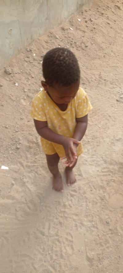 A young child is depicted standing on sandy, uneven ground in Agunmo, Nigeria. The child exhibits dark skin, short, dark, textured hair, and wears a yellow short-sleeved top adorned with white patterns. A small, gold-colored earring is visible in the child's left earlobe. The child stands barefoot, looking downwards, with hands positioned together in front of the body, appearing to be rubbing or clasped. The ground surface consists of light brown sand mixed with small pebbles and dirt. In the upper left background, a light-colored concrete or stucco wall is visible, meeting the sandy terrain at an angle. Scattered white fragments are present on the sand near the base of the wall.