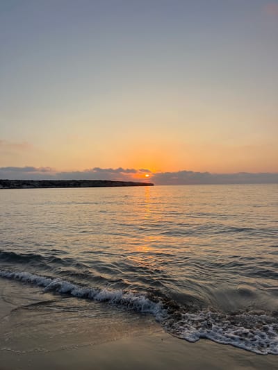 This is a vertical, eye-level, wide shot of a beach at sunset. The image is split roughly in half, with the left side depicting the sky and the right side showing the ocean and sand.

The sky is a gradient of pale blue and orange. The sun, a bright orange orb, is partially obscured by a line of clouds and is located just above the horizon, casting a warm glow. There are some wispy clouds scattered across the sky.

The ocean occupies the right half of the frame. Gentle waves are rolling onto the sandy shore. The water reflects the warm hues of the sunset, creating streaks of gold and orange on its surface. The texture of the water is detailed, with many small ripples. The foam from a receding wave is visible at the edge of the water, creating a frothy line against the sand. The sand itself appears damp and smooth, with subtle textures from the retreating water.

In the distance, along the horizon, a dark, man-made structure, possibly a breakwater or pier, runs horizontally across the image. A small, dark figure, too indistinct to identify as a person or object, is standing on this structure.

The overall mood of the image is serene and peaceful, characteristic of a sunset by the sea. The lighting suggests it is late afternoon or early evening. There is no visible text in the image. The location is Ineia, Cyprus.