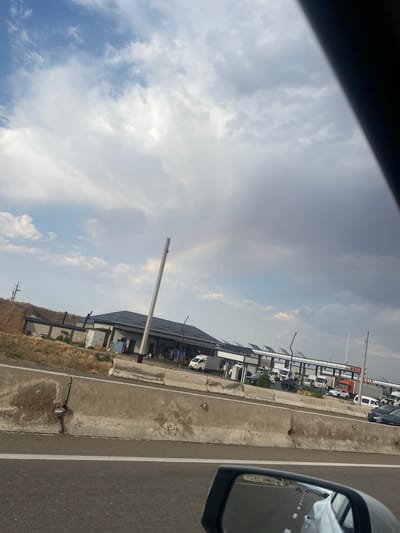 The image depicts a highway scene with a gas station and convenience store complex visible on the right side of the road. The sky is partly cloudy with a faint rainbow visible. Several vehicles, including cars, trucks, and vans, are parked at the gas station. The foreground shows a concrete barrier dividing the highway and the shoulder, with some cracks and damage visible on the barrier. The reflection in the car's side mirror shows the road ahead and other vehicles. The location is identified as being in the city of Kushminor, Uzbekistan. There are no people clearly visible in the image. The environment appears to be a somewhat rural or exurban setting, given the dry grass and undeveloped land visible on the left side of the road. The gas station appears to be operational, with pumps and signage indicating it is a "BENZIN" station, which translates to "gasoline" in Russian and other Turkic languages.