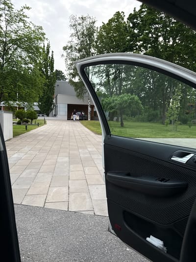 The image depicts a residential driveway in Tabasalu alevik, Estonia. A person is walking away from a dark-colored car towards a modern house. The person is carrying multiple white bags, suggesting they are bringing in groceries or other items. The driveway is paved with light-colored rectangular stones, and bordered by a manicured lawn with some decorative bushes. Lush green trees surround the property. The perspective is from inside a car, with the passenger door open, showing the interior door panel, handle, and window. The sky is overcast.