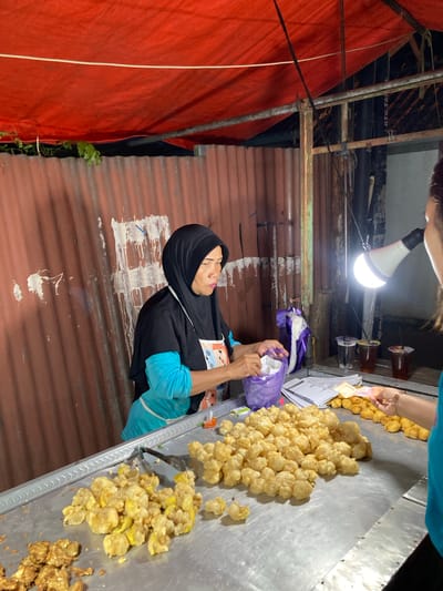 The image shows a woman in a black hijab and blue shirt standing at a food stall in Proyonangg, Indonesia. She is behind a metal counter filled with fried food, possibly snacks or appetizers, in piles. To the left of the counter, a large red tarp is visible. Above the counter, a bright light bulb illuminates the scene, suggesting it might be evening or nighttime. The woman is holding a purple bag and appears to be attending to customers or preparing food. In the background, there are corrugated metal walls, and some construction or makeshift structures are visible. A person's hand with a bracelet is reaching for money and food from the counter. There are cups with dark liquid on a shelf above the counter, and some papers with text are on the counter as well. The overall impression is that of a bustling street food vendor operating at night.