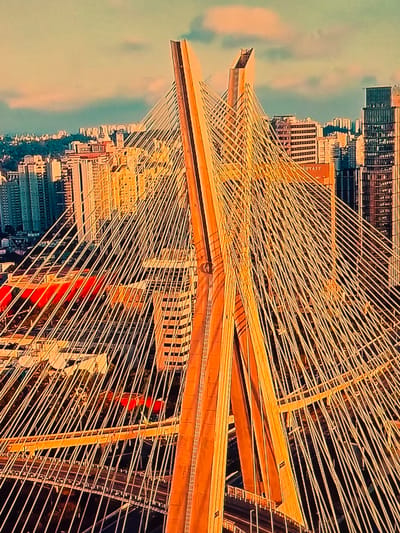 This is an aerial photograph of the Octavio Frias de Oliveira Bridge in São Paulo, Brazil, at sunset. The bridge, a cable-stayed structure, is the central focus of the image. Its two distinctive orange towers rise vertically, supporting a dense network of white cables that fan out across the scene. The golden light of the setting sun illuminates the bridge and the surrounding urban landscape. In the background, a dense cityscape unfolds, characterized by numerous tall buildings and high-rise structures. The sky above is a gradient of pale orange and soft blue, with wispy clouds. The lower portion of the image reveals the multi-layered roadway of the bridge, with traffic lanes visible. The overall impression is one of architectural grandeur and the vibrant energy of a major metropolis.