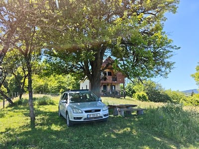 A silver Ford Focus is parked on a grassy area next to a wooden picnic table and benches. Behind the car, a multi-story wooden house with balconies and a pitched roof is visible, partially obscured by a large, leafy tree. The scene is set in a natural environment with lush green grass and trees, suggesting a rural or mountainous location. In the background, more trees and hills under a clear blue sky indicate a sunny day, likely in the late morning or early afternoon. The overall impression is one of a peaceful, outdoor setting, possibly a vacation spot or a secluded residence. The license plate on the car reads "XR-300-BA".