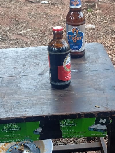 The image depicts two bottled beverages placed on a weathered wooden table in an outdoor setting in Egede, Nigeria. In the foreground, slightly left of center, is an uncapped, dark brown glass bottle of Amstel Malt, distinguished by its red label and partially visible "NO ALCOHOL" text. To its right and slightly behind it, a brown glass bottle of Tiger Lager Beer, with a blue and silver label featuring a tiger logo, stands upright. The wooden table displays visible grain and signs of wear. Underneath the table's edge, a green promotional banner for "glo text 4 millions Super Promo" offering "N555M WORTH OF PRIZES" is partially visible. Directly below the table, a white bowl containing remnants of food and a metal spoon is present. The background shows dry, reddish-brown earth with scattered leaves and small debris, indicative of an unpaved outdoor area. No individuals are visible in the scene.