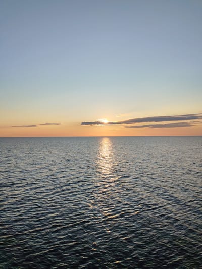 The image depicts a calm sea at sunset. The sun, partially obscured by a thin layer of clouds, casts a golden glow across the water, creating a shimmering reflection. The sky transitions from a pale blue at the top to a warm orange near the horizon. The sea is a deep blue, with small ripples disturbing its surface. The scene is serene and natural, with no visible human presence or man-made structures. The location, Kakkarovo, Russia, is not visually evident in the image.