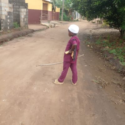 A young child, estimated to be between 3-6 years old, stands on a dirt road in Ijotan, Nigeria, facing slightly away from the viewer towards the left. The child wears a white skullcap, a long-sleeved maroon top, matching maroon trousers, and light-colored slip-on sandals. Their left hand is placed on their hip, while their right hand holds a dark object, possibly a fan, and a light-colored object, possibly a piece of fruit or another small item. A light-colored wooden stick lies on the dirt road in front of the child.
The road is unpaved, consisting of reddish-brown earth with some lighter patches. To the left of the road, in the mid-ground, there are partially constructed block walls, possibly a fence or building foundation, with some green vegetation growing in a narrow trench or drainage channel alongside the road. Further back on the left, a light yellow building with a dark brown base and a small wooden gate is visible.
To the right of the road, the ground is darker and appears muddy or damp, with sparse green vegetation and some debris. Trees with green foliage line the right side of the road in the background, casting some shadows. The sky is not visible.