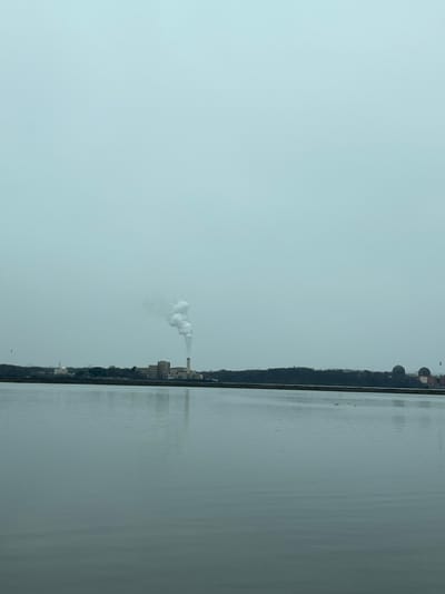 The image depicts a wide-angle view across a calm body of water, likely the Hudson River, towards a distant shoreline in the Town of Cortlandt, United States of America, under an overcast sky. The foreground is dominated by the gray, reflective surface of the water, extending approximately two-thirds up the frame. The horizon features a low-lying landmass populated by industrial structures and trees.

On the left side of the distant shore, a large industrial facility is visible, characterized by multiple buildings and a tall, slender smokestack from which a dense, white plume of smoke or steam ascends vertically before dissipating into the pale sky. To the far right, partially obscured by a dark tree line, are two prominent, light-colored dome-shaped structures, typical of nuclear reactor containment buildings, associated with the Indian Point Energy Center. Adjacent to these domes are additional industrial buildings. The sky is uniformly light gray or muted blue, devoid of clear features. Two faint, dark specks, possibly birds, are visible in the sky, one on the far left and another on the far right. No explicit human figures or immediate interactions are discernible. The overall environment suggests a cool, possibly hazy or foggy, day.