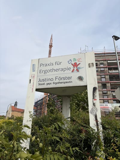 The image shows a sign for "Praxis für Ergotherapie Justina Förster" (Practice for Occupational Therapy Justina Förster) in Würzburg, Germany. The sign is white with black text and a red and blue logo depicting stylized figures. Behind the sign, a multi-story building is under construction, covered in scaffolding. A construction crane is visible in the sky. Green bushes and trees are in the foreground. A street lamp is to the right of the sign. The sky is overcast. The sign itself has some graffiti on it, including a circular sticker with the word "FUCK" and a shield with an emblem. Another sticker, a red shield with white markings, is below the graffiti. A banner on the construction site reads "WORK ON PROGRESS". The building appears to be located on a street, with another building visible in the background to the left, which has a distinctive red roof and a spire.