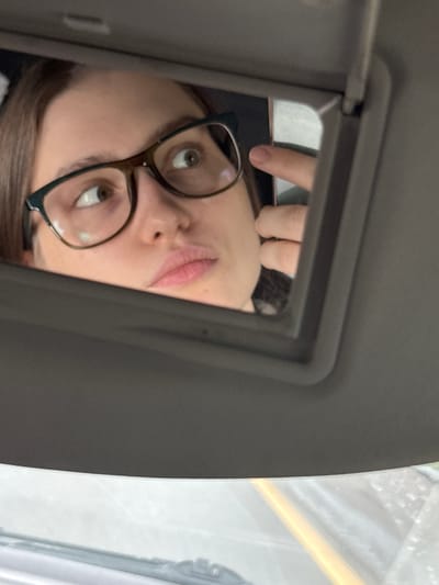 The image depicts a person with brown hair and glasses reflected in a car's sun visor mirror. The person's face is partially obscured by the mirror frame and their own finger. Outside the car window, the background is blurred, obscuring detail. The image does not provide any information regarding the location in Myaglovo, Russia.