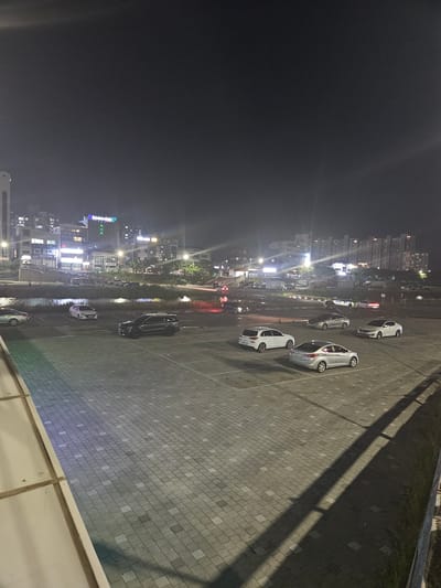 The image depicts a nighttime scene in Eumnae-ri, South Korea, featuring a parking lot adjacent to a body of water and a city skyline. The parking lot is paved with square tiles and contains several parked cars.
In the foreground, a light-colored wall or railing is visible on the left. The parking lot is mostly empty, with a few cars parked in designated spots. A dark-colored SUV is parked prominently in the middle right of the frame. To its left, a white sedan is parked. Further back, another white sedan and a silver sedan are visible. On the far left, a silver sedan is parked.
Beyond the parking lot, a narrow body of water separates it from the opposite bank. Along the opposite bank, there are trees and vegetation. Further inland, a cityscape is visible with various buildings. Some buildings have illuminated windows and signs, suggesting they are commercial or residential structures. A prominent sign with blue and green lights is visible on one of the buildings. Streetlights are also present, illuminating the area. The sky is dark, indicating it is nighttime.
The overall impression is a quiet, urban scene at night, with the parking lot serving as a central element of the composition, framed by the natural element of the water and the man-made environment of the city. There are no people visible in the image.