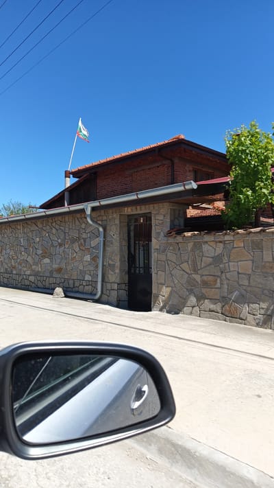 A street view in Boliartsi, Bulgaria, shows a stone wall bordering a property with a brick house visible above. A Bulgarian flag flies from the house’s chimney. The wall has a metal gate. A car's side mirror is visible in the foreground. The sky is clear and blue.