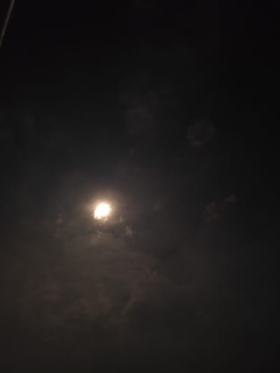 The image displays a nocturnal scene featuring a brightly illuminated moon partially veiled by clouds. The moon, positioned towards the left-center of the frame, emits an intense white-yellow glow, creating a radiant halo effect through the translucent, light grey clouds immediately surrounding it. These clouds are visibly lit by the moon's light, standing out against the dark, deep grey to black night sky.
Scattered across the rest of the frame are darker, less-illuminated cloud formations, indicating significant cloud cover. A faint, thin, diagonal line, possibly a wire or a distant structure, is visible in the upper-left corner of the image. Another extremely subtle, wavy line can be discerned in the bottom-right quadrant.
The scene captures a cloudy night, with the moon as the primary source of light, lending a serene and atmospheric quality. There are no visible people, distinct man-made objects (apart from the faint lines), or discernible text. The setting is the night sky above Lhokmambang, Indonesia.
