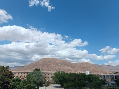 This image depicts a scenic view of Balkanabat City, Turkmenistan, under a bright blue sky dotted with fluffy white clouds. The foreground is dominated by lush green trees, some of which are tall and mature, casting shadows on the ground. Partially visible behind the trees are several buildings, appearing to be residential or administrative structures, with multiple windows and satellite dishes.
In the mid-ground, a large, imposing mountain range with a reddish-brown hue rises majestically. The mountains exhibit a textured surface, suggesting a rugged, natural landscape. Several smaller hills and mountain formations are visible in the distance, fading into a hazy horizon.
Among the buildings, a modern, slender white tower stands out, contrasting with the older structures and the natural surroundings. The presence of apartment-style buildings and the satellite dishes suggests a settled urban or semi-urban environment.
The overall impression is one of a clear, sunny day, possibly late morning or early afternoon, given the bright lighting and the absence of long shadows. The scene evokes a sense of tranquility and the harmonious coexistence of urban development and the natural landscape. There is no visible text in the image.