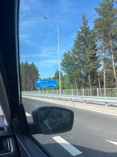 An exterior, eye-level, medium shot captures a road sign on a highway in Volchikhinskii Maidan, Russia, under a bright blue sky with scattered white clouds. The sign, predominantly blue with white text and arrows, indicates directions to "ЧЕРНУХА" (Chernukha) with a left-pointing arrow and "ВОЛЧИХИНСКИЙ МАЙДАН" (Volchikhinskii Maidan) with a right-pointing arrow. The road itself is asphalt, with white lane markings visible. To the right of the road, a metal guardrail separates the highway from a sandy embankment, beyond which a dense forest of pine trees is visible. A single lamppost stands tall on the left side of the road, its light fixture angled towards the highway. The image is framed by the interior of a vehicle, with a dark-colored car door and a side mirror reflecting the road and sky. The overall scene suggests a clear day on a rural highway.