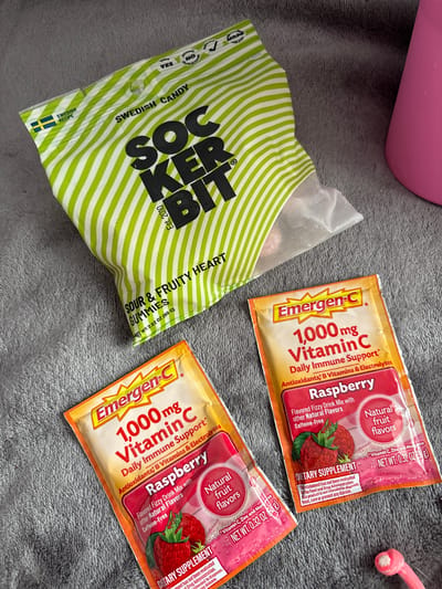 The image shows a gray surface, possibly a blanket or rug, on which several items are placed. There's a green and white striped bag of "Sockerbit" Swedish candy. Next to it are two packets of "Emergen-C" vitamin C drink mix in raspberry flavor. The packets are orange with red accents and display information about the product. The bag of candy is labeled "Sour & Fruity Heart Gummies". A pink object and a pink object with white details, are also visible. The scene does not provide enough information to specify a location in Seal Beach, United States of America.
