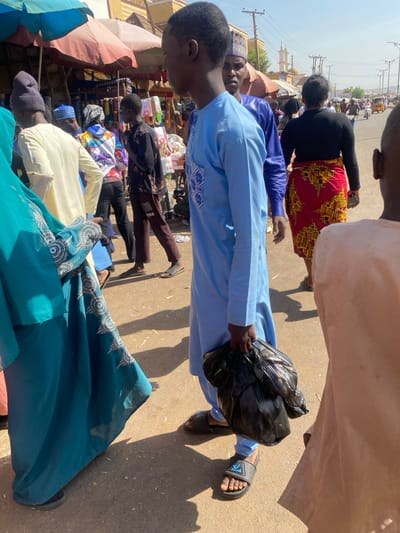 This image depicts a bustling outdoor street market scene in Sabon Gari South, Nigeria, under bright daylight. The primary subject is a young adult male, of dark complexion, positioned centrally, wearing a light blue long-sleeved traditional garment (likely a kaftan) with white embroidery on the left chest. He wears black sandals with a white logo and holds a large, lumpy black plastic bag in his left hand, hanging at his side. He is looking towards the viewer's left.
To his immediate left, a portion of a female figure is visible, wearing a teal-colored abaya or long dress with intricate silver or white embroidery along the hem and sleeves. Black flat footwear is visible on her left foot.
Behind the central male, another adult male with dark complexion is partially visible, wearing a dark blue traditional long-sleeved garment and a matching blue embroidered cap. He looks generally forward.
On the right side of the frame, another adult female is seen from behind, walking away. She wears a black long-sleeved top and a vibrant red skirt with a repeating yellow and black geometric pattern. A portion of another individual in a light tan garment is visible on the far right.
In the background, numerous individuals move through the market. Visible figures include a person in a cream-colored garment and a dark knitted cap, another in a multicolored hooded sweatshirt, and a person in a dark shirt and brown pants. Several market stalls with striped fabric awnings (red and blue/teal) are evident, displaying various goods. The ground is a mix of dirt and uneven pavement, casting strong shadows from the individuals. In the far distance, a street or road with vehicles, power lines, and poles can be seen. A light-colored building with a distinctive architectural feature, possibly a mosque, stands on the distant horizon.