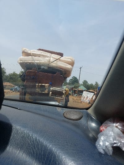 The image depicts an outdoor scene in Hoss, Nigeria, viewed from the interior of a vehicle. The primary subject is a dark-colored truck, positioned ahead on an unpaved, reddish-brown road. The truck is overloaded with household goods, specifically a tall stack of light-colored mattresses, individually wrapped in clear plastic and secured with dark straps or ropes. Beneath the mattresses are several darker, rectangular furniture components, likely disassembled sofa frames. A single individual, potentially a female, wearing a yellow headscarf and darker clothing, stands near the truck's rear on the right side. The background features a clear blue sky, scattered green foliage, and several rudimentary buildings or market stalls constructed from earth-toned materials and corrugated metal roofs. A utility pole with power lines is visible on the right. A large, partially obscured white banner with colorful graphics stands in the background to the far right. The immediate foreground shows a dark, textured vehicle dashboard with a circular speaker grille and a clear plastic bag containing red items on the far right.
