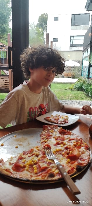 The image, taken on 27/02/2020 at 10:25, depicts a young male seated at a wooden table in a dining area in Tumbaco, Ecuador. The male, with dark, curly hair, wears a light-colored t-shirt featuring a Spiderman graphic and text. He is positioned left of center, looking towards the camera with a slight facial expression. In the foreground, a round metal pizza pan contains a partially eaten pizza topped with pepperoni, cheese, and corn kernels. A fork with a wooden handle rests on the pizza. A single slice of this pizza is on a white rectangular plate to the male's right. The background, visible through a large window or opening, shows an outdoor garden area with green grass, plants, and a light-colored bench. A multi-story white building with dark-framed windows is visible in the distant background, along with trees and additional vegetation. A dark, possibly wooden, structure is present to the far left, suggesting a playground or outdoor seating feature.
