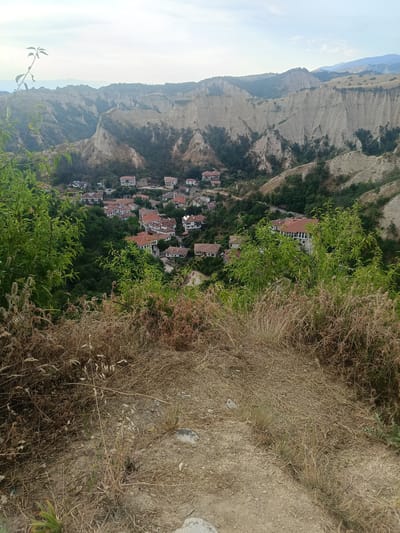 This media file depicts a scenic view of Melnik, Bulgaria, a town nestled within a valley of unique sand pyramids. The shot is taken from an elevated position, looking down upon a collection of traditional houses with red-tiled roofs and white walls, scattered amongst lush green trees.

In the foreground, dry, golden grasses and some green foliage frame the scene, suggesting a natural, somewhat wild environment. The background is dominated by the striking geological formations of the sand pyramids, their eroded shapes creating a dramatic and distinctive landscape. These geological features rise steeply, with pockets of vegetation clinging to their slopes.

The time of day appears to be late afternoon or early evening, as indicated by the soft, diffused light and the slightly hazy sky. The weather seems calm, with no strong winds or rain visible. The overall mood conveyed by the image is one of serene beauty and historical charm, highlighting the picturesque setting of Melnik and its unique natural surroundings. There is no visible text in the image.