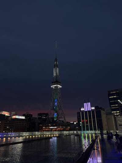 The image depicts a night scene in Nagoya, Japan, likely at the Oasis 21 complex. A tall, lattice tower, identifiable as the Nagoya TV Tower, dominates the center of the frame, illuminated with blue and green lights along its structure. In the foreground, a reflective water surface, part of Oasis 21's "Water Spaces," mirrors the surrounding lights and the twilight sky, which shows hues of deep blue transitioning to a subtle pinkish-red near the horizon. Several buildings are visible to the left and right of the tower. To the right, a modern building features illuminated vertical lines of colored lights and a purple illuminated sign reading "ESTHE TWIN". People are present, walking along the elevated walkway and near the edge of the water feature, their forms partially silhouetted against the illuminated environment. The overall atmosphere is urban and modern, with a focus on illuminated architecture and public spaces at dusk.