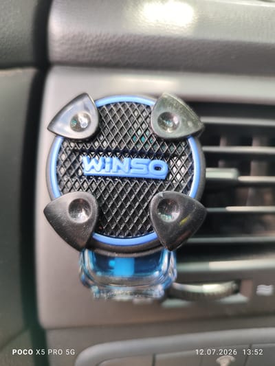 A car air freshener with the brand name "WINSO" is clipped onto a car's air vent. The air freshener is round with a textured black surface and a blue trim. Four triangular black pieces are attached to the outer edge of the air freshener, each with a small reflective element. A blue liquid is visible in a transparent container below the air freshener. The background shows a grey car interior, including the air vent slats. The image is timestamped "12.07.2026 13:52" and branded "POCO X5 PRO 5G". The image was taken in Lypivka, Ukraine.