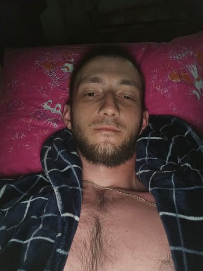 A man with a beard lies on a pink pillow with a bird pattern, wearing a dark blue robe with white checks. A thin silver chain is visible on his chest, which has dark hair. The lighting is dim. The image does not contain any information about the city of Serebriansk, Kazakhstan.