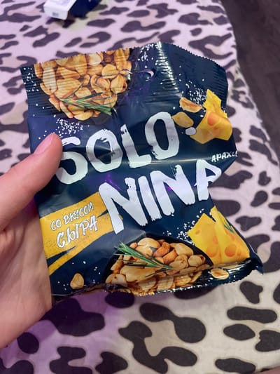 A hand is holding a crumpled dark blue snack bag with images of peanuts, cheese, and rosemary. The bag has white text that reads "SOLO NINA" and "СО ВКУСОМ СЫРА" (with cheese flavor). The background is a patterned blanket or fabric. The image appears to be taken indoors. There is no information about Vaganovo, Russia in the image.