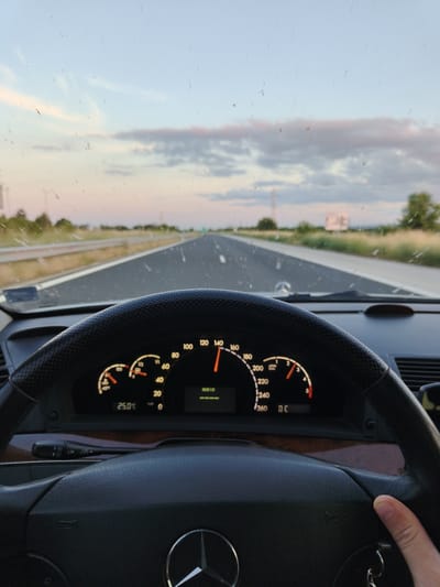 A first-person perspective shot from inside a car on a highway at dusk. The car's dashboard is visible, showing a speedometer indicating a speed around 140 km/h, an odometer reading 25.0 C, and a tachometer. The steering wheel, partially in the foreground, features a Mercedes-Benz logo. The road stretches ahead under a cloudy sky with hues of pink and blue. The windshield has some dust or water spots. To the left and right of the road, grassy fields and trees are visible, with a distant billboard on the right. The location is identified as Momkovo, Bulgaria, suggesting the highway is near this area.