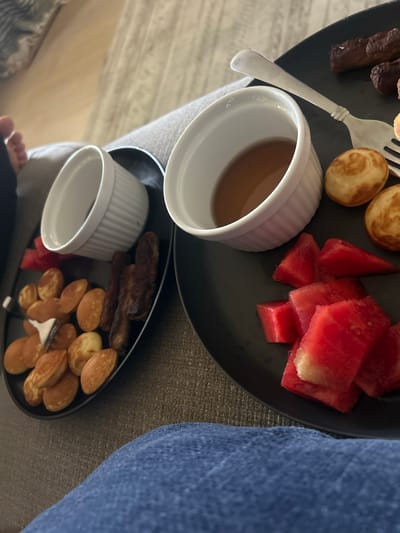 This image shows two black plates on what appears to be a person's lap, possibly in a home setting. The first plate contains what looks like cooked sausages, small round pancakes or "puffs," watermelon pieces, a fork, and a white ramekin filled with a brown liquid, likely syrup. The second plate also contains sausages, pancakes, watermelon, a fork and a white ramekin, but it appears to be empty. The person is wearing what appears to be denim clothing. The image provides no information to determine if the location is in Reno, USA.