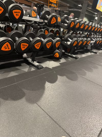Rows of black dumbbells with orange labeling, resting on metal racks, dominate the left side of the image. The dumbbells are labeled with various weights, including 16, 18, 20, 22, and 26. The brand names "ZIVA" and "MATRIX" are visible on some of the dumbbells and racks. A portion of the gym floor is visible on the right. Shadows cast by the dumbbells and racks are prominent on the floor.