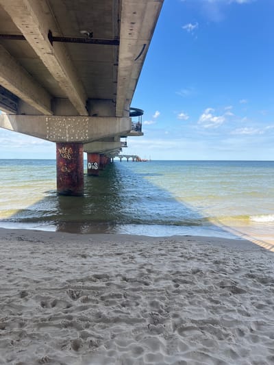 This is a ground-level, eye-level, wide shot of the Międzyzdroje Pier in Międzyzdroje, Poland, on a sunny day. The pier's concrete structure and supports dominate the left and center of the frame, casting a shadow on the beach and water. The supports are rusted and covered in graffiti, with the words "WIG" and "OIS" visible on one of them. Waves gently lap the sandy shore in the foreground, and the calm sea extends to the horizon under a bright blue sky with scattered white clouds. In the distance, the pier continues, with a few distant figures visible near its end. The scene is one of a quiet, pleasant day at the beach.